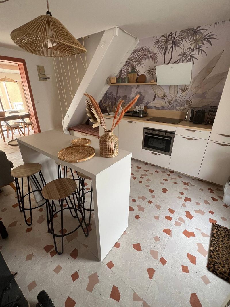 Pièce de vie : îlot de cuisine, suspension en rotin, papier peint palmiers et sol terrazzo